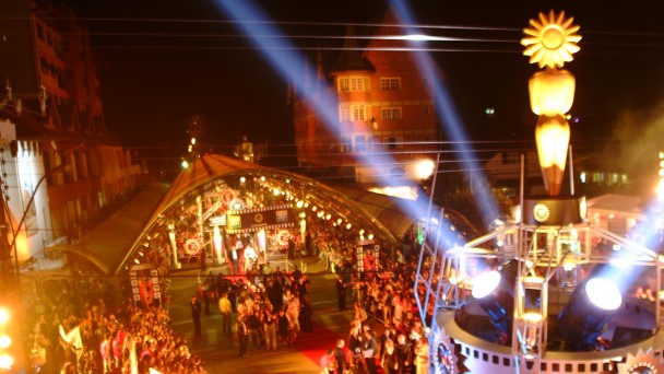 Vista de cima da entrada do festival de cinema de Gramado, com p&uacute;blico aglomerado em frente ao tapete vermelho. Um grande Kikito iluminado aparece em destaque no primeiro plano
