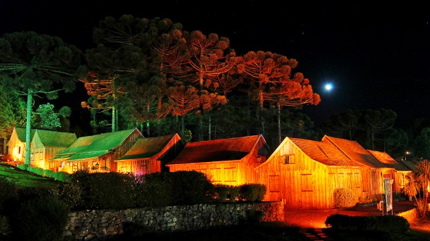 Casas t&iacute;picas de madeira da serra ga&uacute;cha iluminadas &agrave; noite, sob as arauc&aacute;rias, em noite de lua cheia.
