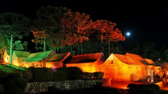 Casas t&iacute;picas de madeira da serra ga&uacute;cha iluminadas &agrave; noite, sob as arauc&aacute;rias, em noite de lua cheia.